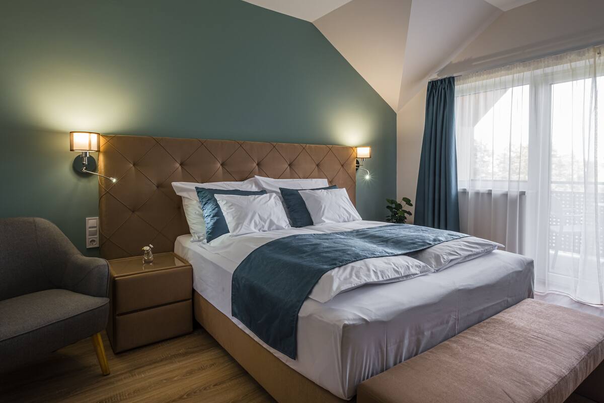A modern hotel room with a tufted headboard, a large bed with white and blue bedding, a bedside table with a lamp, a grey armchair, and a window with curtains.
