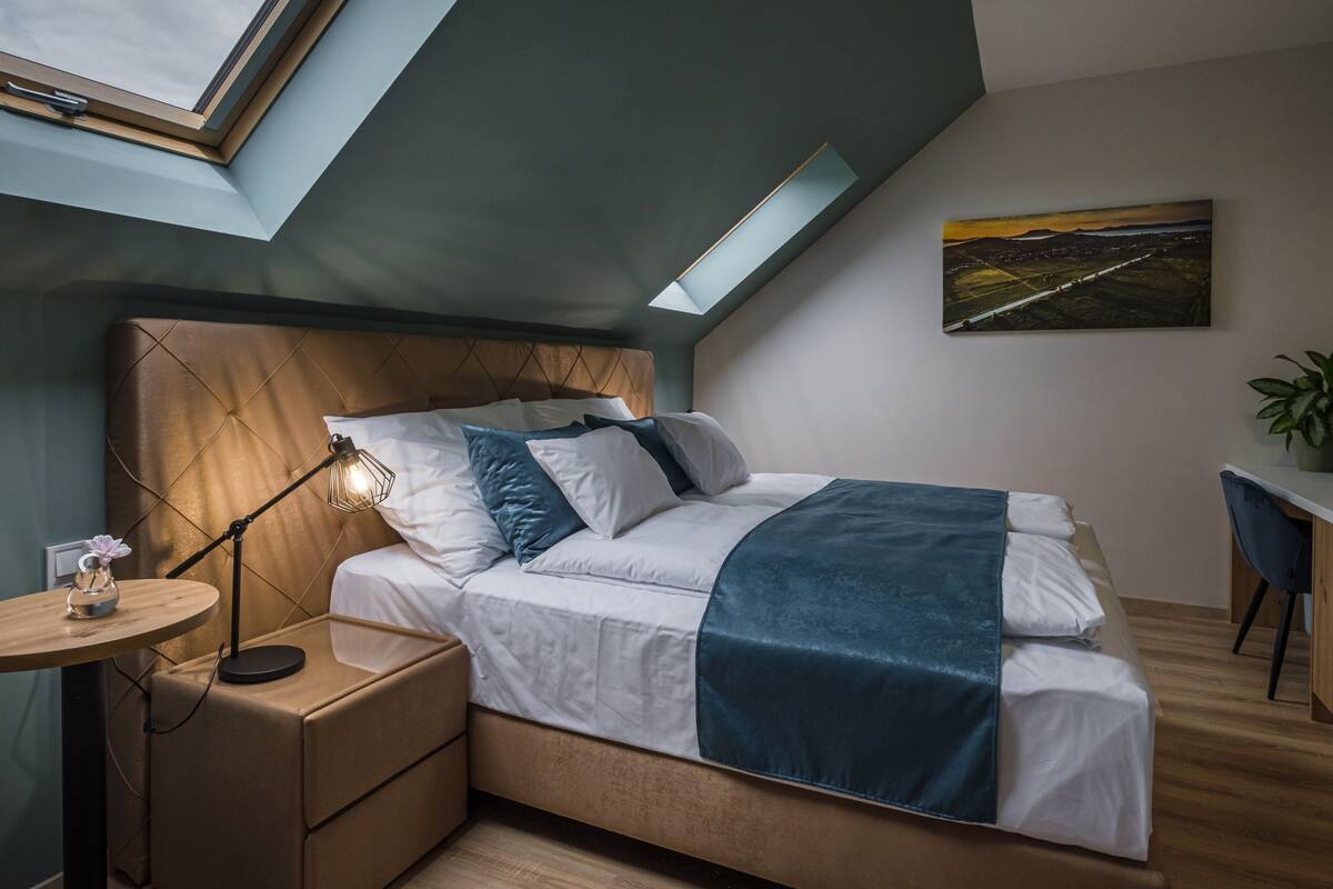 A bedroom with a skylight, a bed with a blue blanket, a bedside table with a lamp, and a painting on the wall.
