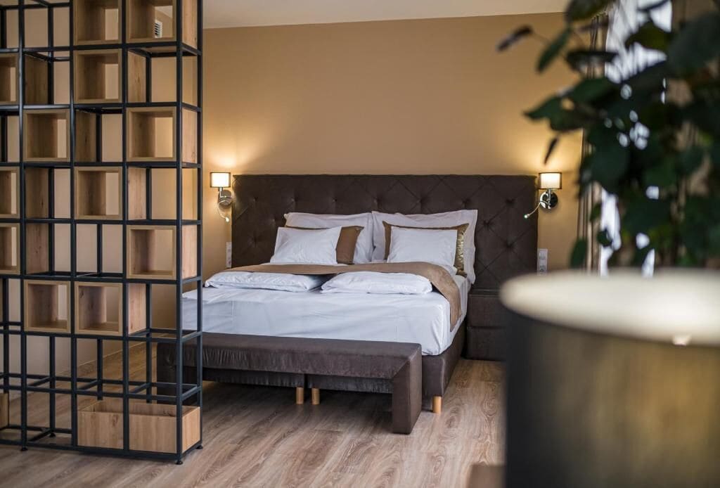 Un dormitorio con cama, cabecera, lámparas de noche, suelo de madera y una planta.