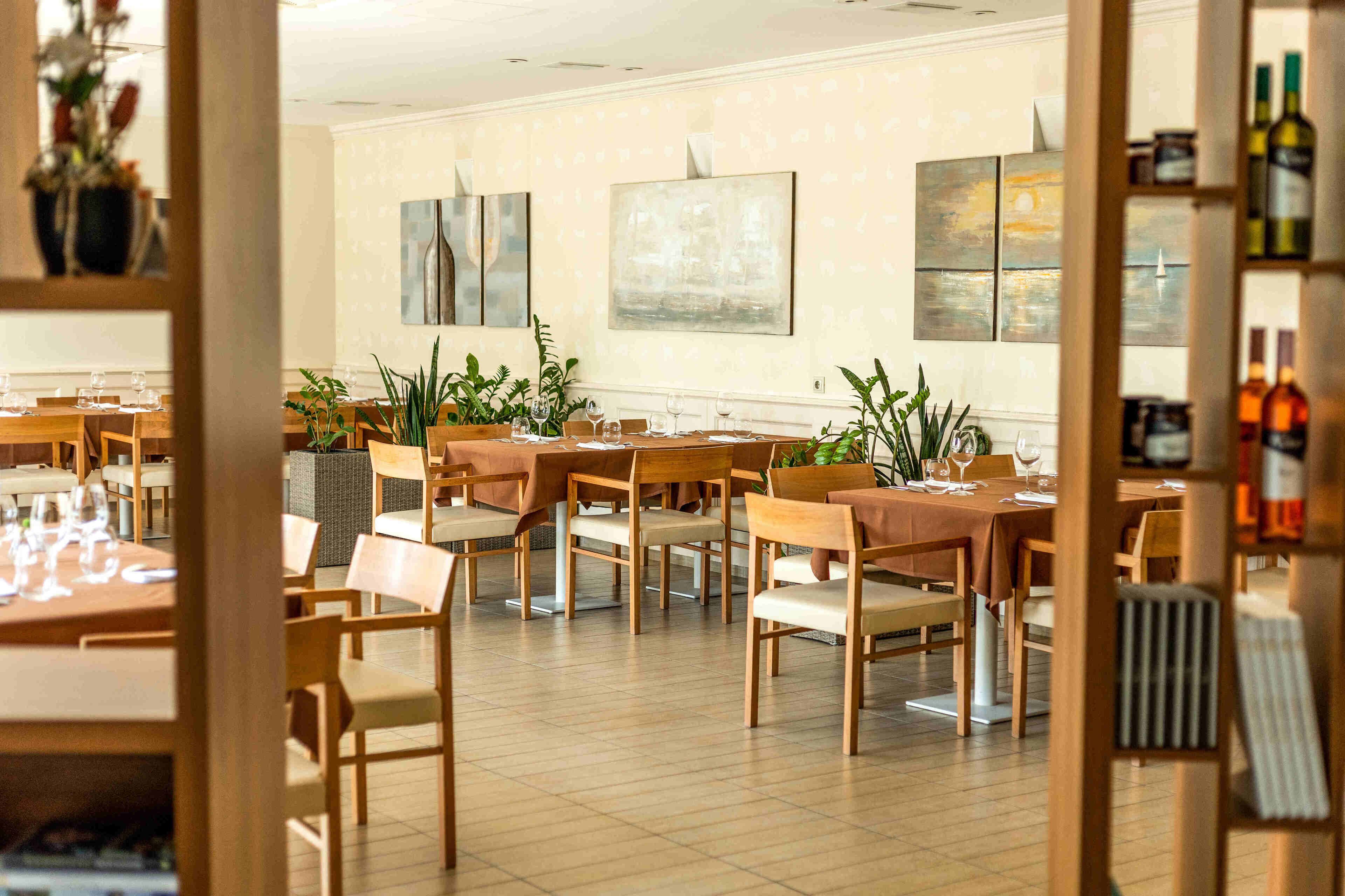 Restaurante com várias mesas e cadeiras, algumas com plantas. Existem obras de arte emolduradas nas paredes.