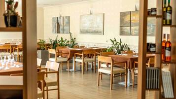 Restaurante com várias mesas e cadeiras, algumas com plantas. Existem obras de arte emolduradas nas paredes.