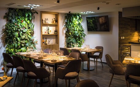 A dining area with two tables set for six, surrounded by chairs. There are vertical gardens on the walls, a wooden shelf with bottles, and a television mounted on the wall.