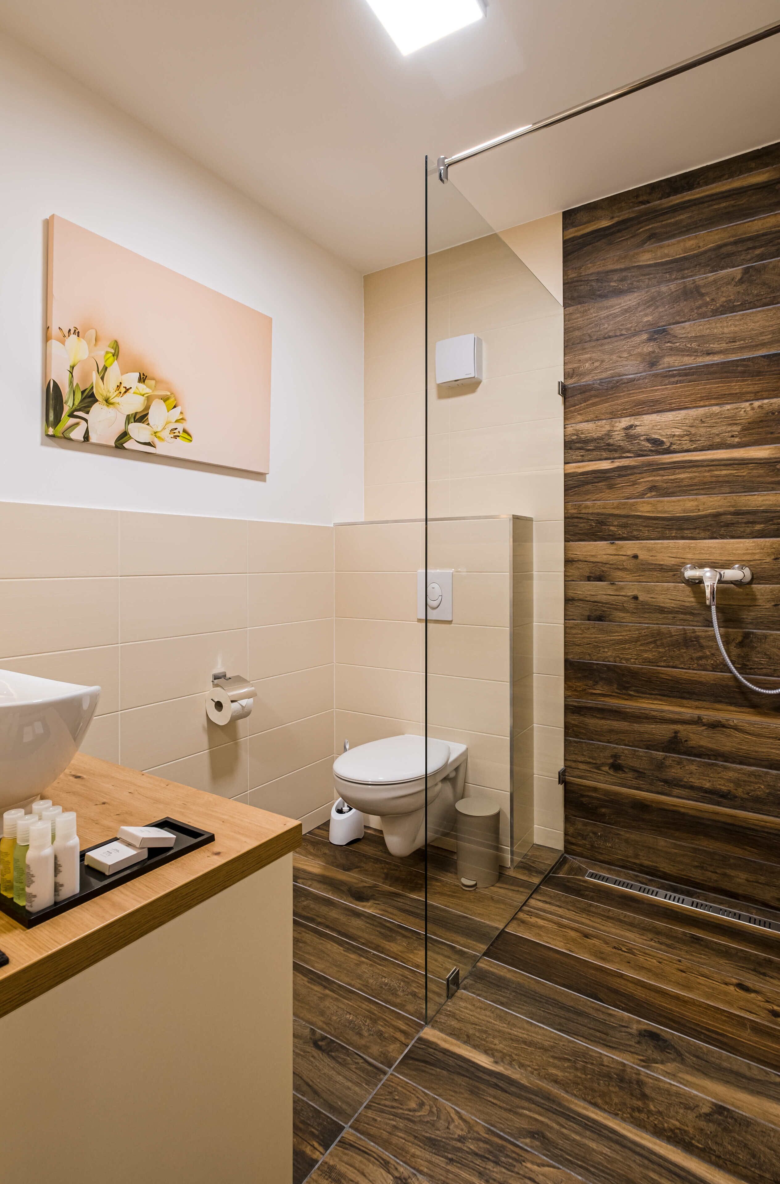 A modern bathroom with a wooden floor, a glass shower enclosure, a toilet, and a sink with toiletries.