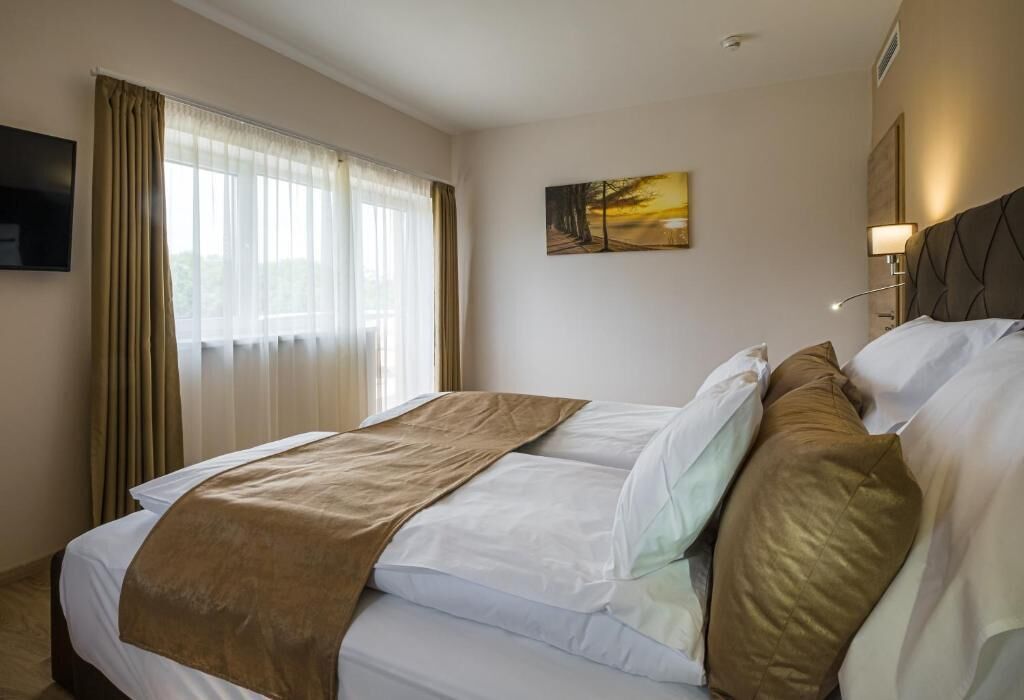 Een hotelkamer met een bed, gordijnen, een schilderij en een nachtkastje met een lamp.