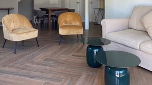 Een moderne woonkamer met een visgraatparketvloer, twee leren fauteuils, een ronde glazen salontafel, een beige bank en een eethoek met een tafel en stoelen.