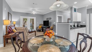 A dining area with a glass table, chairs, and a vase of flowers. There is a ceiling fan, a television, and a kitchen in the background. - Elegant 3-bedroom condo with 2 King beds/2 twins close to OWA/Foley Sports Plex. (Foley)