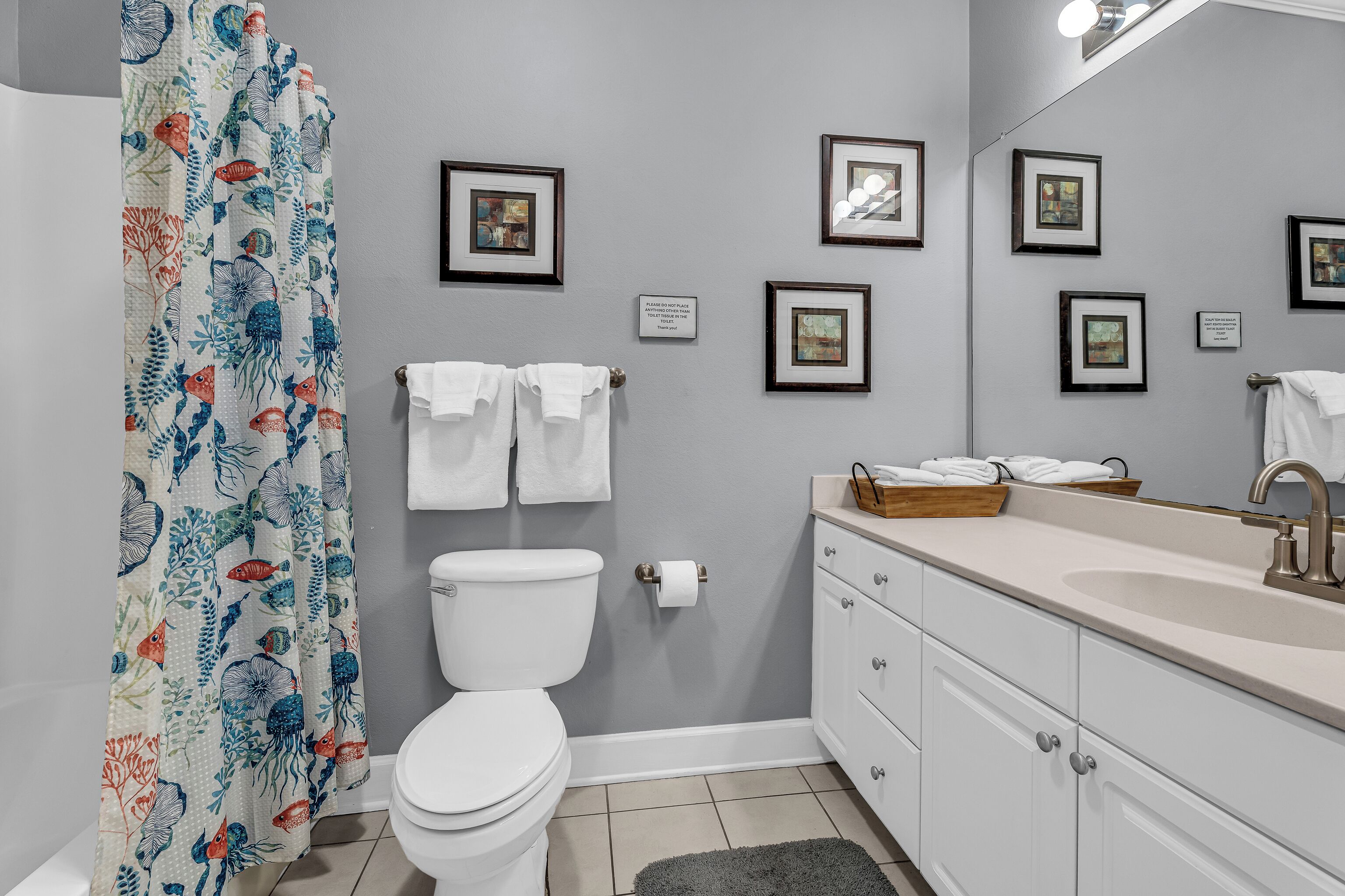 A bathroom with a toilet, sink, and shower curtain with a fish pattern. There are framed pictures on the wall and towels hanging on the wall.