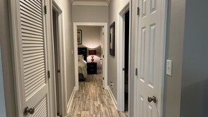 A hallway with a wooden floor, white doors, and a bed with a nightstand in the background. - Elegant 3-bedroom condo with 2 King beds/2 twins close to OWA/Foley Sports Plex. (Foley)