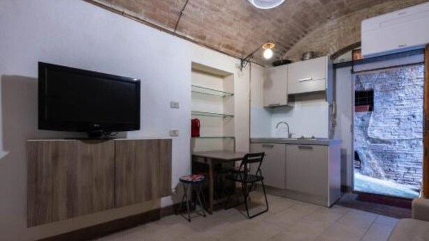 A compact kitchen with a dining area, a television on the wall, and a stone wall feature. - Schöne, Kleine Wohnung in Zentraler Lage (Siena)