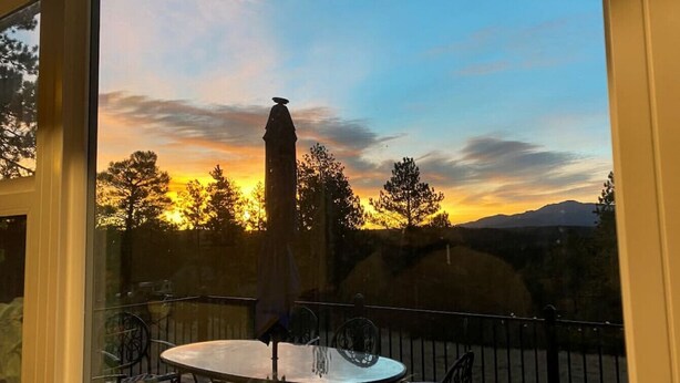 A deck with a round table and chairs, overlooking a sunset with trees and mountains in the background. - 24 Quiver Circle · Spectacular Mountain View ★ 5 Acres ★ PIKES PEAK (Florissant)