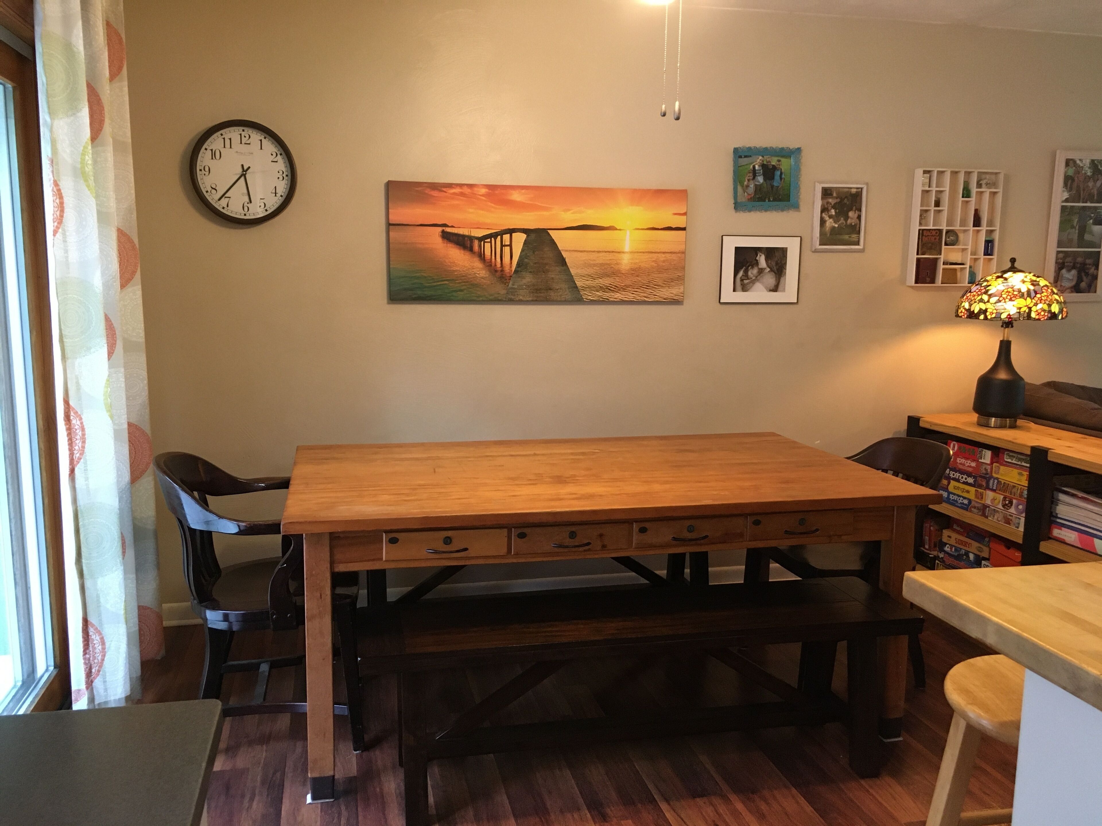 A dining area with a wooden table, chairs, a bench, a clock, and a painting of a sunset over a bridge.