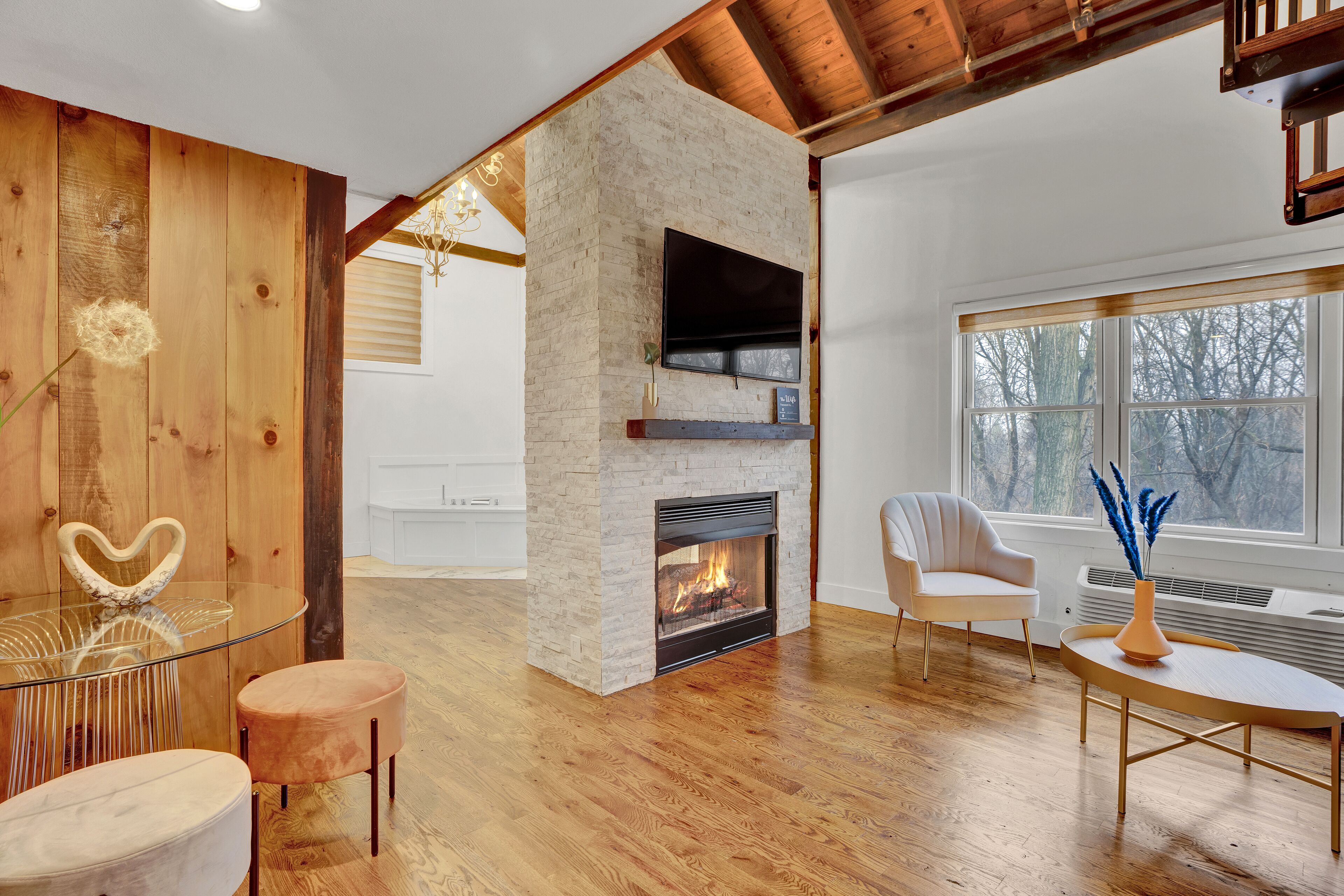 A modern living room with a stone fireplace, wooden floors, and a large window with a view of trees.