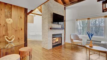 A modern living room with a stone fireplace, wooden floors, and a large window with a view of trees.