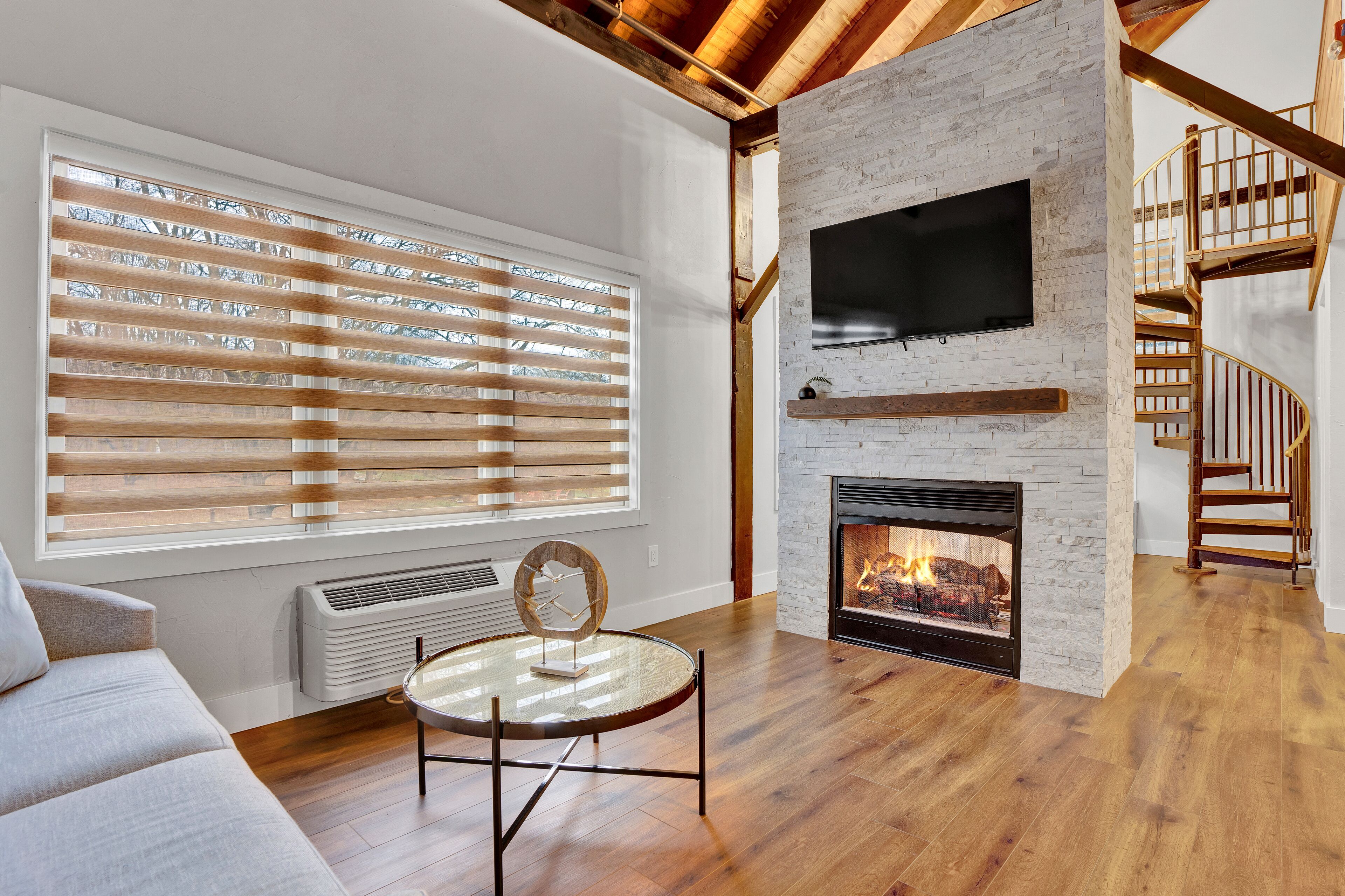 A modern living room with a fireplace, wooden flooring, a round glass table, and a staircase with wooden railings.