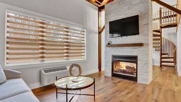 A modern living room with a fireplace, wooden flooring, a round glass table, and a staircase with wooden railings.