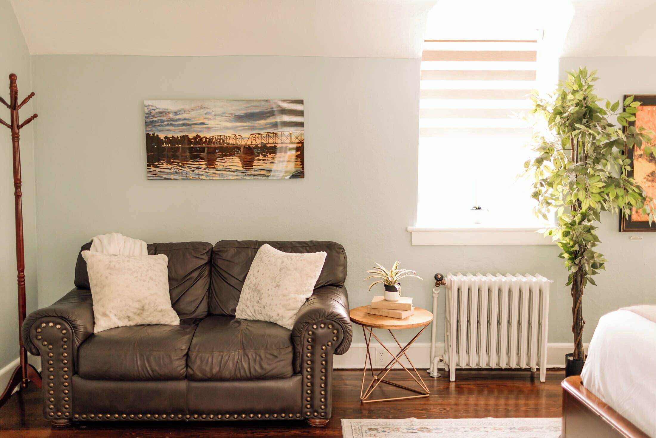 Um quarto aconchegante com um sofá de couro escuro, uma pintura de paisagem urbana, um radiador, piso de madeira e um vaso de plantas.