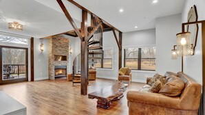 A spacious living room with a stone fireplace, wooden flooring, a leather sofa, and a staircase with exposed beams.