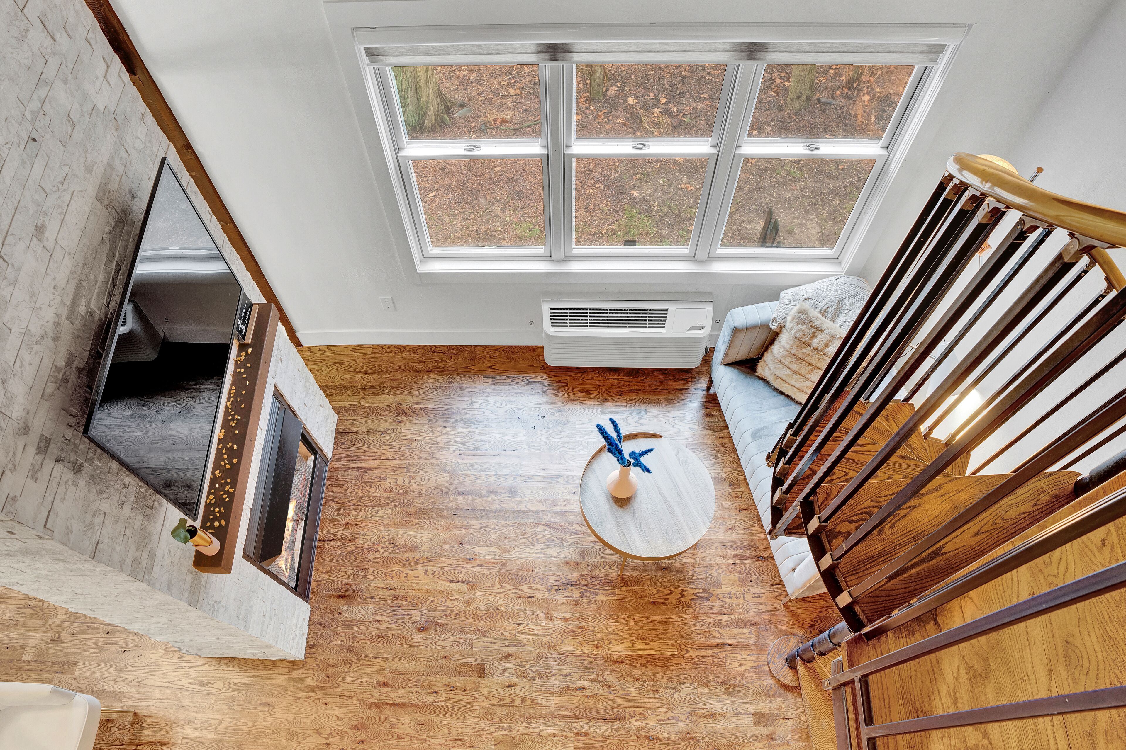 A spacious room with a wooden floor, a large window with a view of trees, a round wooden table, and a staircase with wooden railings.