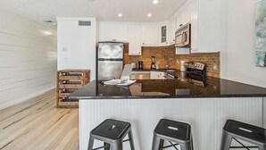 Eine moderne Küche mit weißen Schränken, einem Edelstahl-Kühlschrank, einer Mikrowelle und einer Wandverkleidung aus Backsteinen. Vor der Arbeitsplatte befinden sich drei Barhocker.