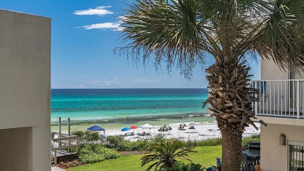 Blick auf einen Strand von einem Balkon aus, mit einer Palme, Strandsonnenschirmen und blauem Himmel.