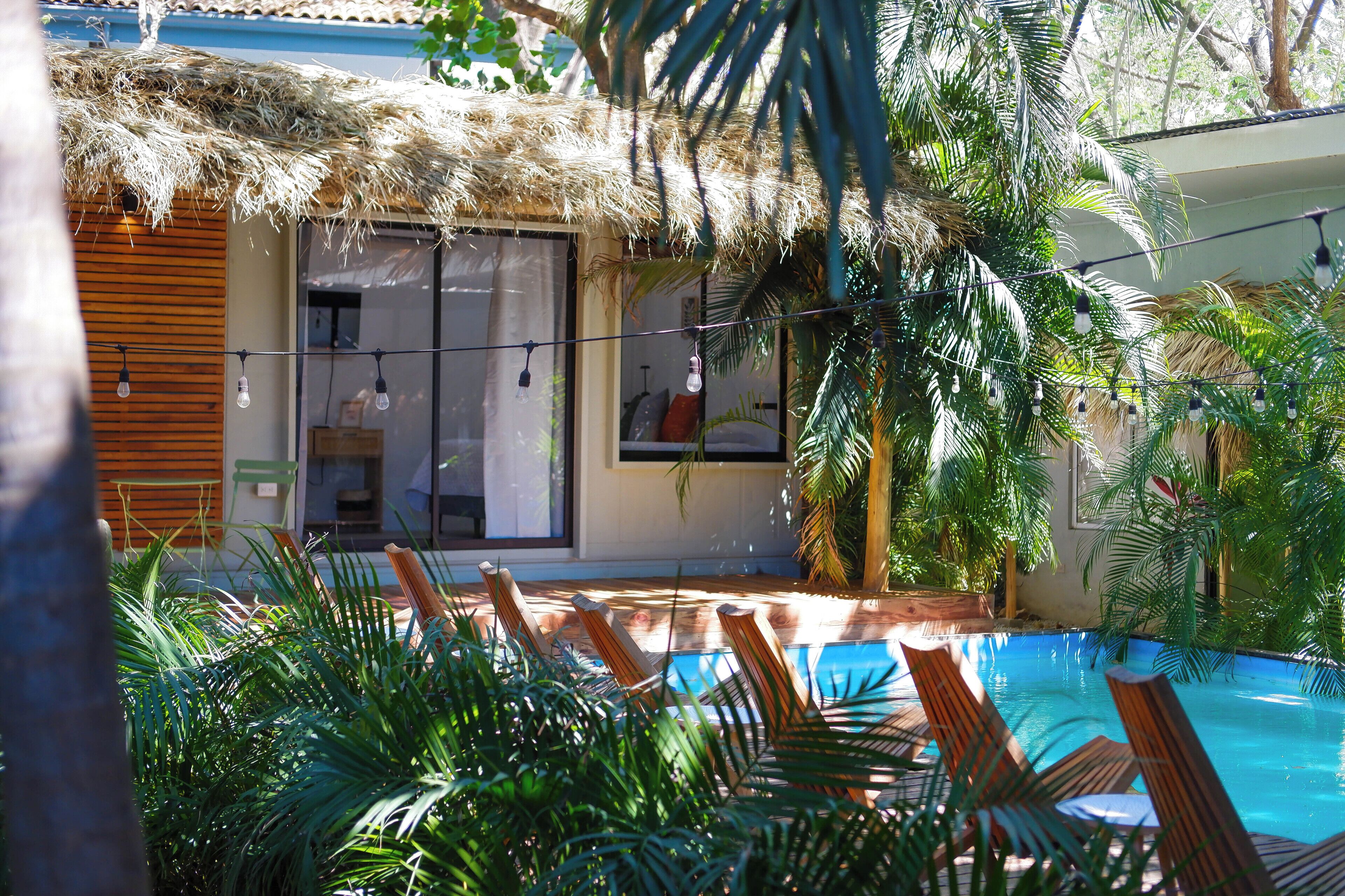 A pool area with wooden loungers, a thatched roof structure, and lush greenery.