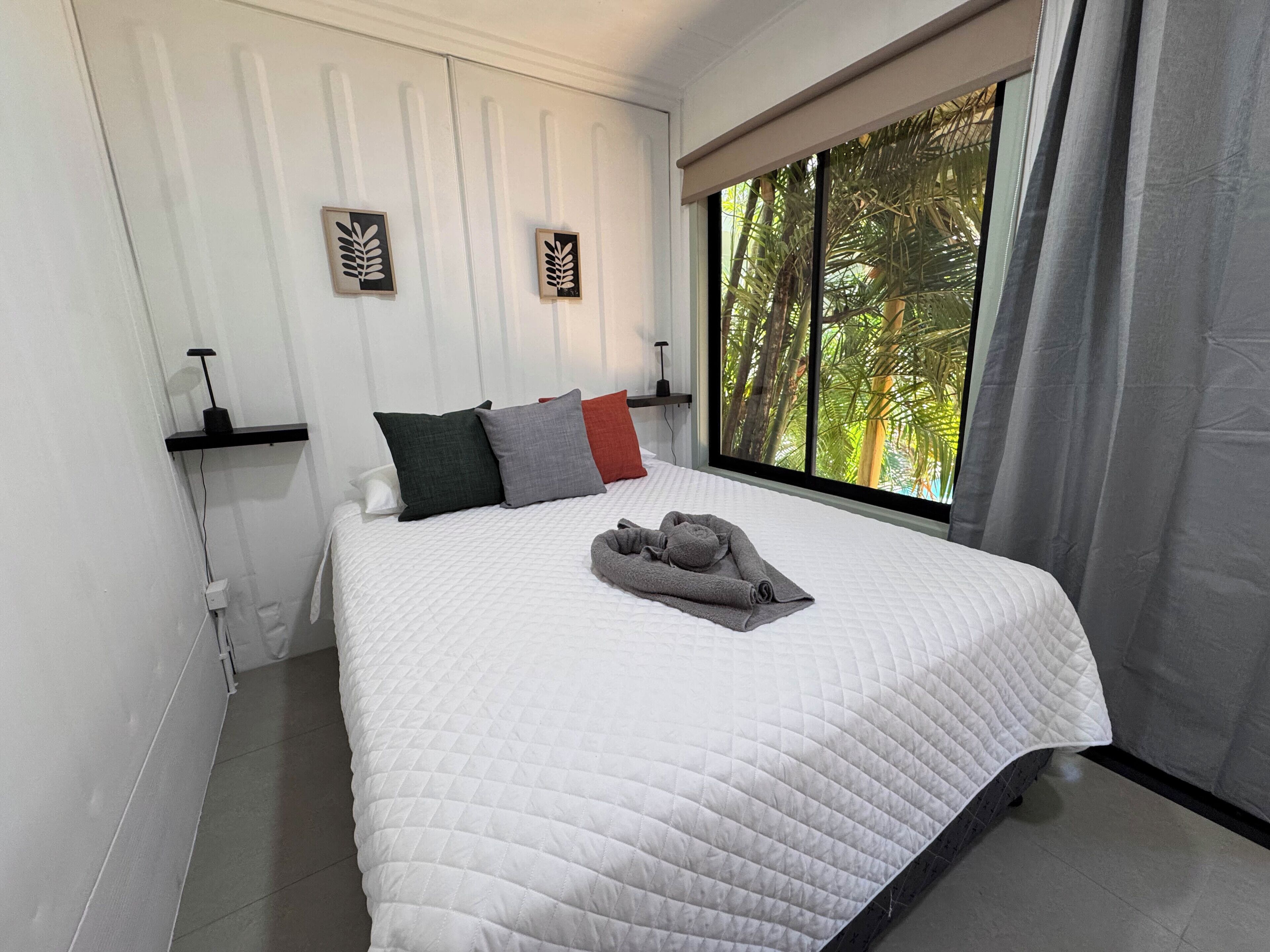 A neatly made bed with a white quilt, pillows in various colors, and a rolled towel on top. There are two wall-mounted light fixtures and a window with a view of palm trees.