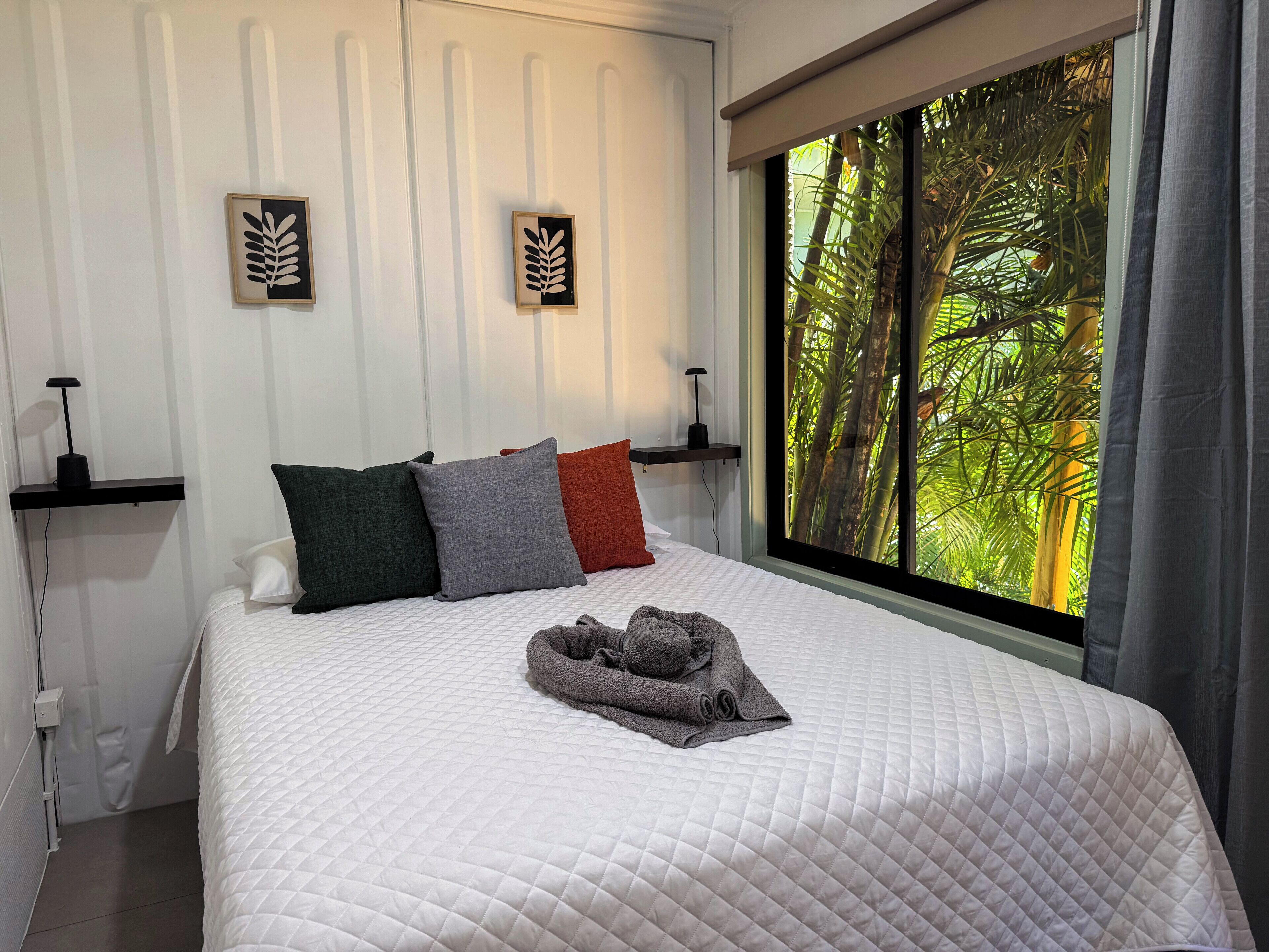 A neatly made bed with a quilted white duvet, a grey towel, and three pillows in green, grey, and red. There are two wall-mounted picture frames with leaf designs. A large window with a view of green foliage is visible.