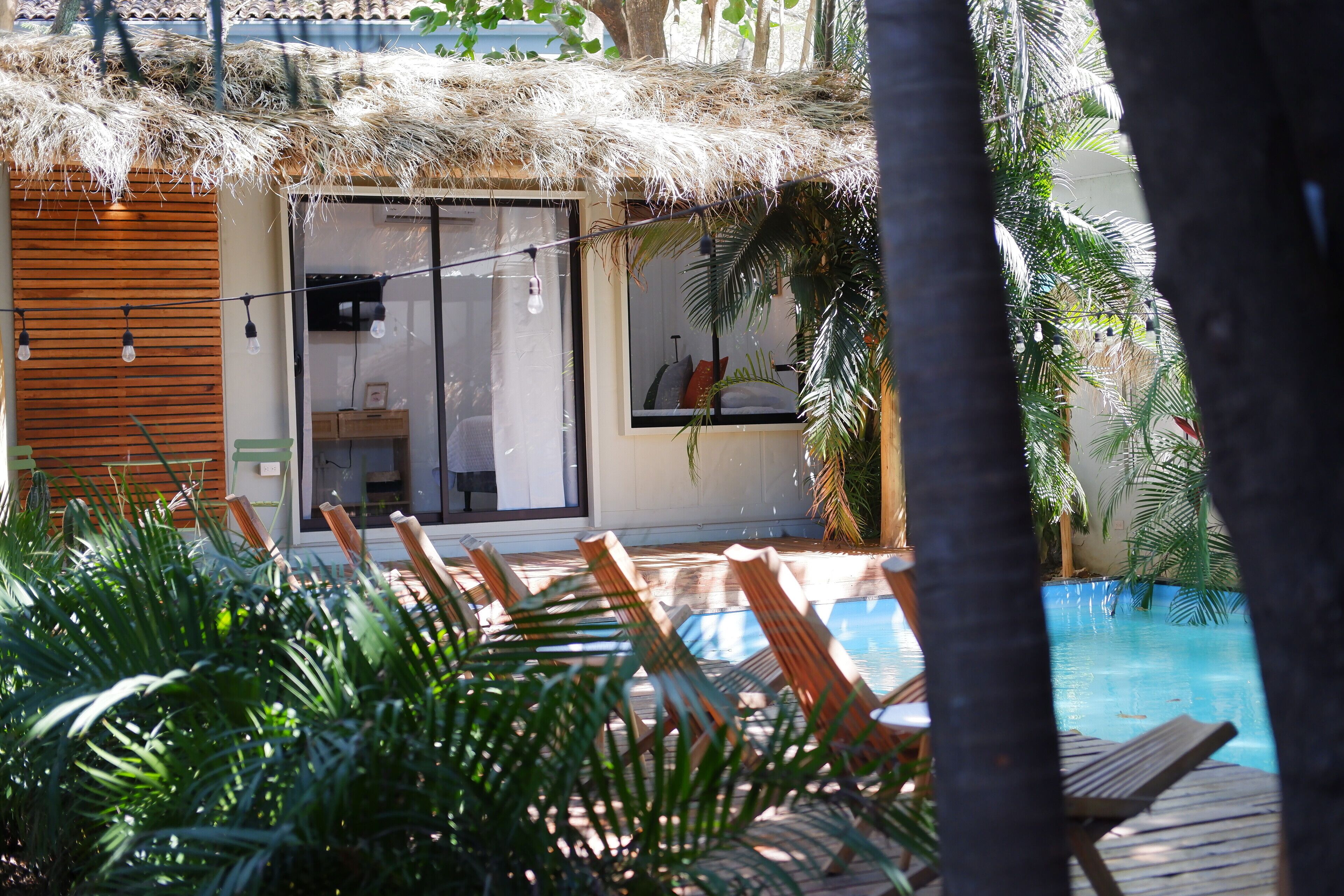 A poolside area with wooden loungers, a thatched-roof structure, and a view of a building with glass doors.
