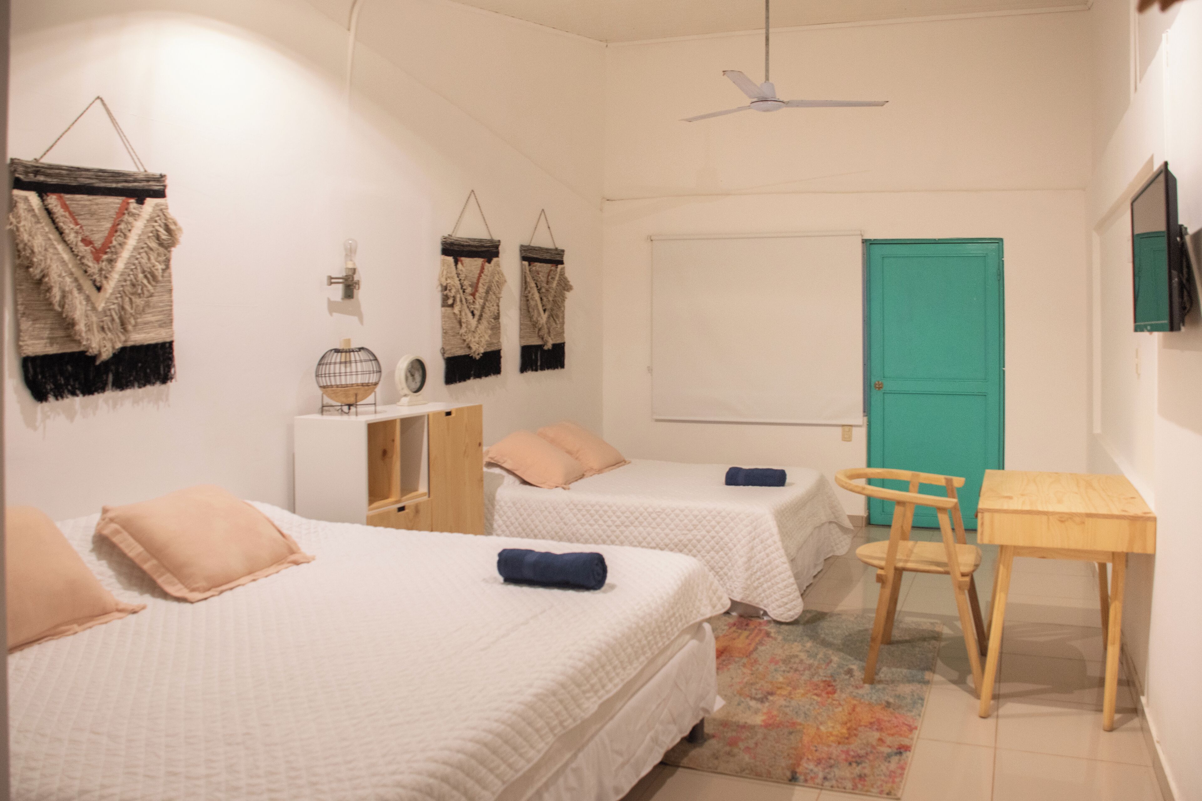 Een kleine, schone kamer met twee bedden, een bureau, een stoel, een blauwgroene deur en een plafondventilator.