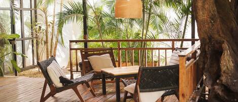 A wooden deck with outdoor furniture, surrounded by lush tropical plants and a large tree trunk.