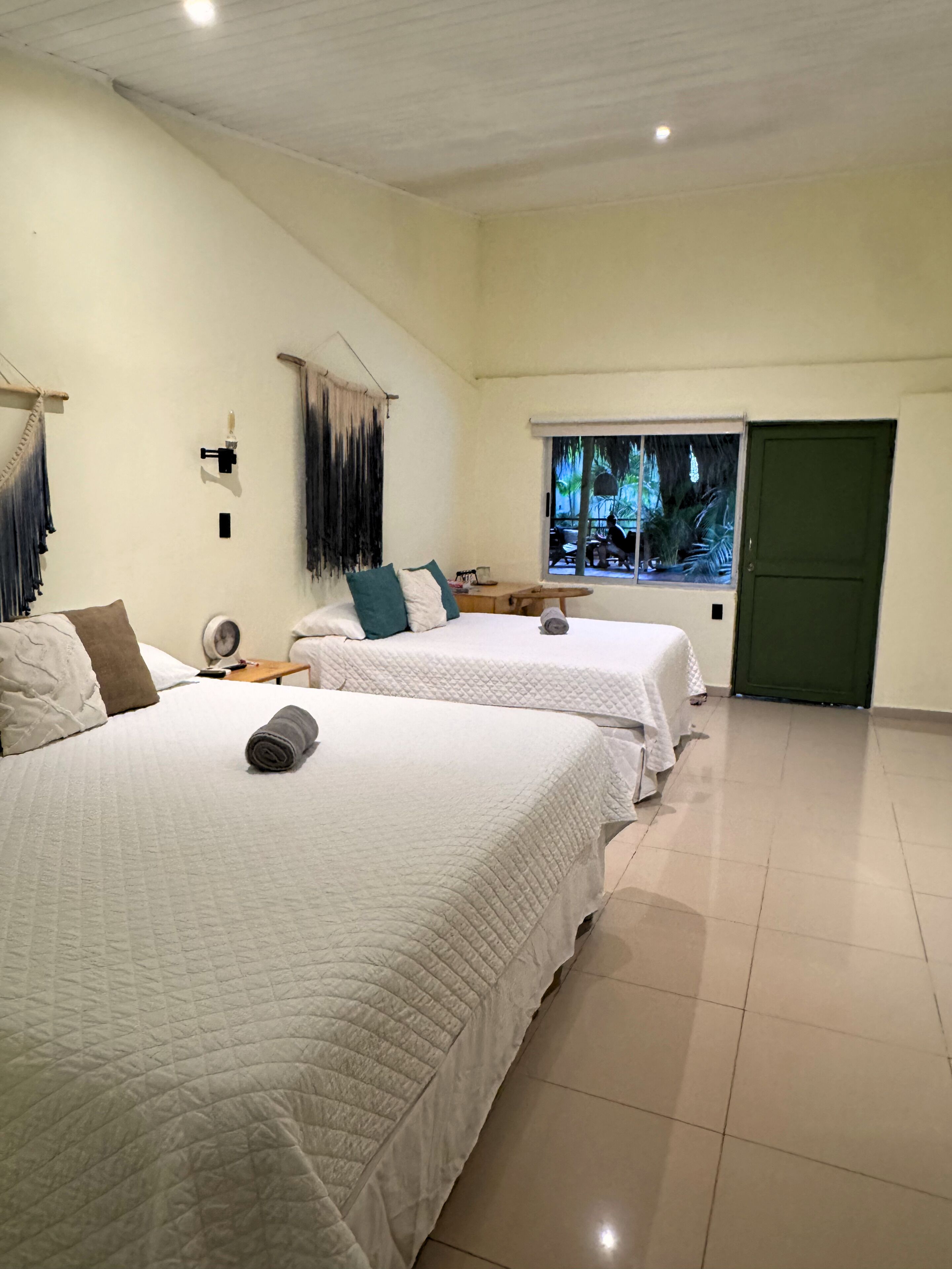 A hotel room with two beds, a small table, a chair, a window with a view of palm trees, and a green door.