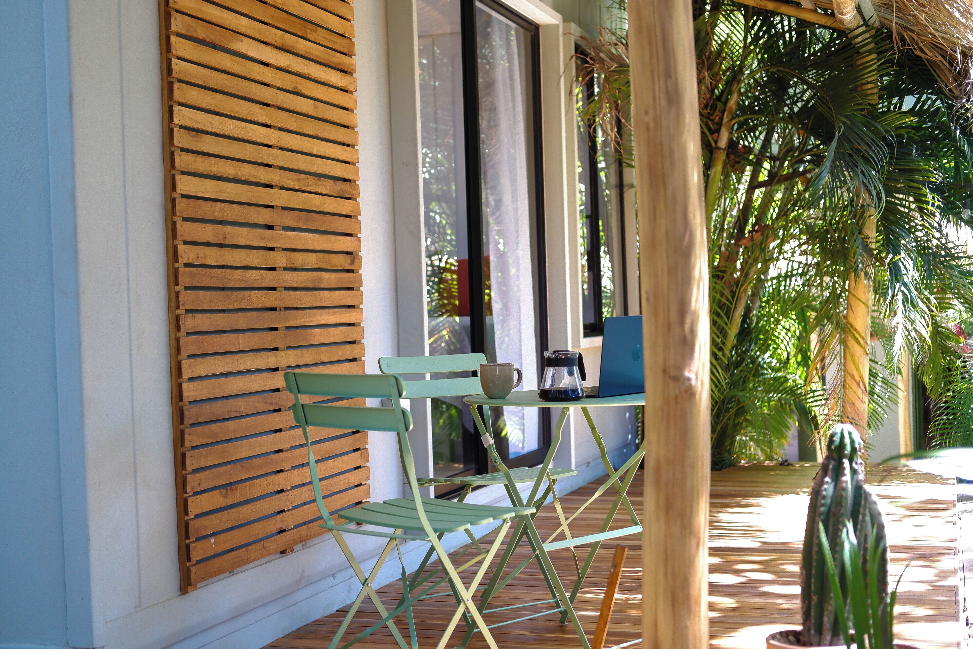 Una ventana con marco de madera y persianas horizontales, una silla plegable verde, una mesita con una cafetera y una planta en maceta.