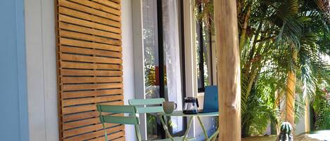 Una ventana con marco de madera y persianas horizontales, una silla plegable verde, una mesita con una cafetera y una planta en maceta.