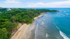 A coastal area with a sandy beach, dense green vegetation, and a few buildings in the background. - Refugio Lodge (Tamarindo)