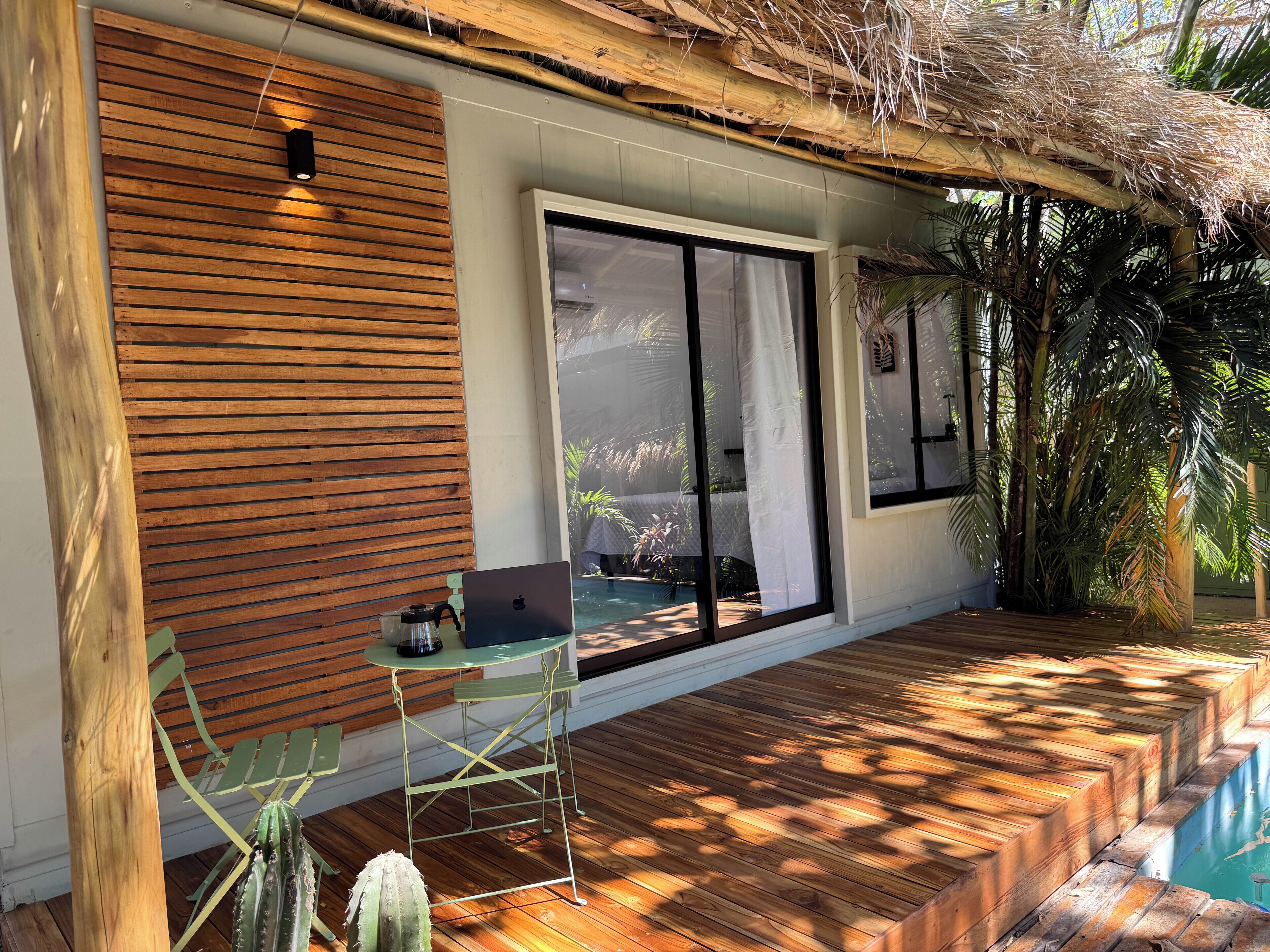 Une terrasse en bois avec un ordinateur portable posé sur une table, un cactus et une piscine.