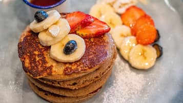 Ein Stapel Pfannkuchen mit Bananenscheiben, Erdbeeren und Blaubeeren, dazu eine kleine Schüssel Ahornsirup.