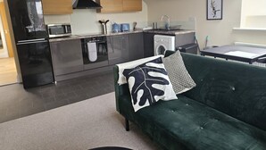 A modern kitchen with a built-in oven, microwave, and dishwasher. There is a green sofa with a decorative pillow and a small table with a chair. - 1 Bed Flat - Sleeps 2 - Parking - Wifi (Newport)