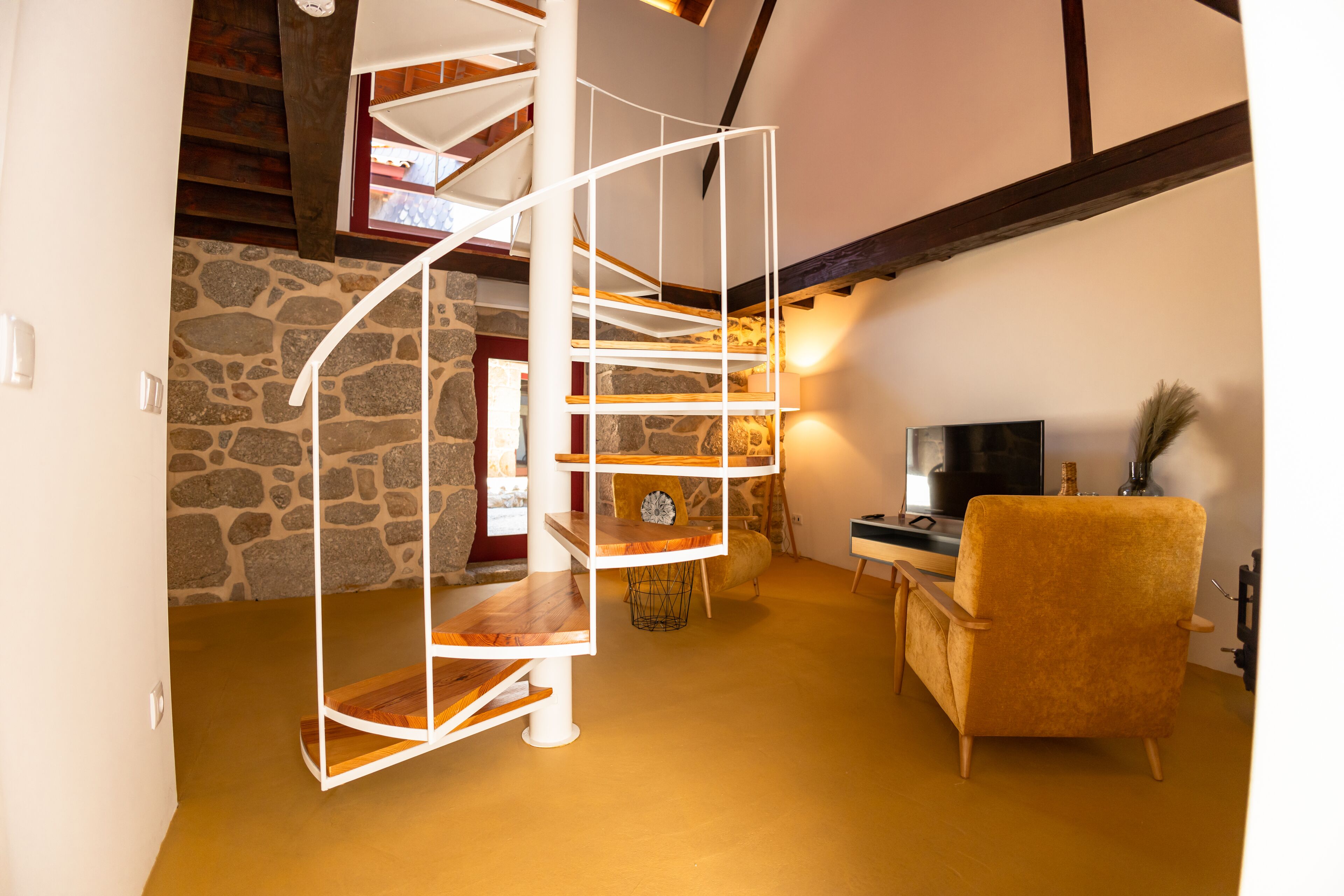 A spiral staircase with wooden steps and white railings, a stone wall, a yellow armchair, and a television in a cozy room with a warm ambiance.