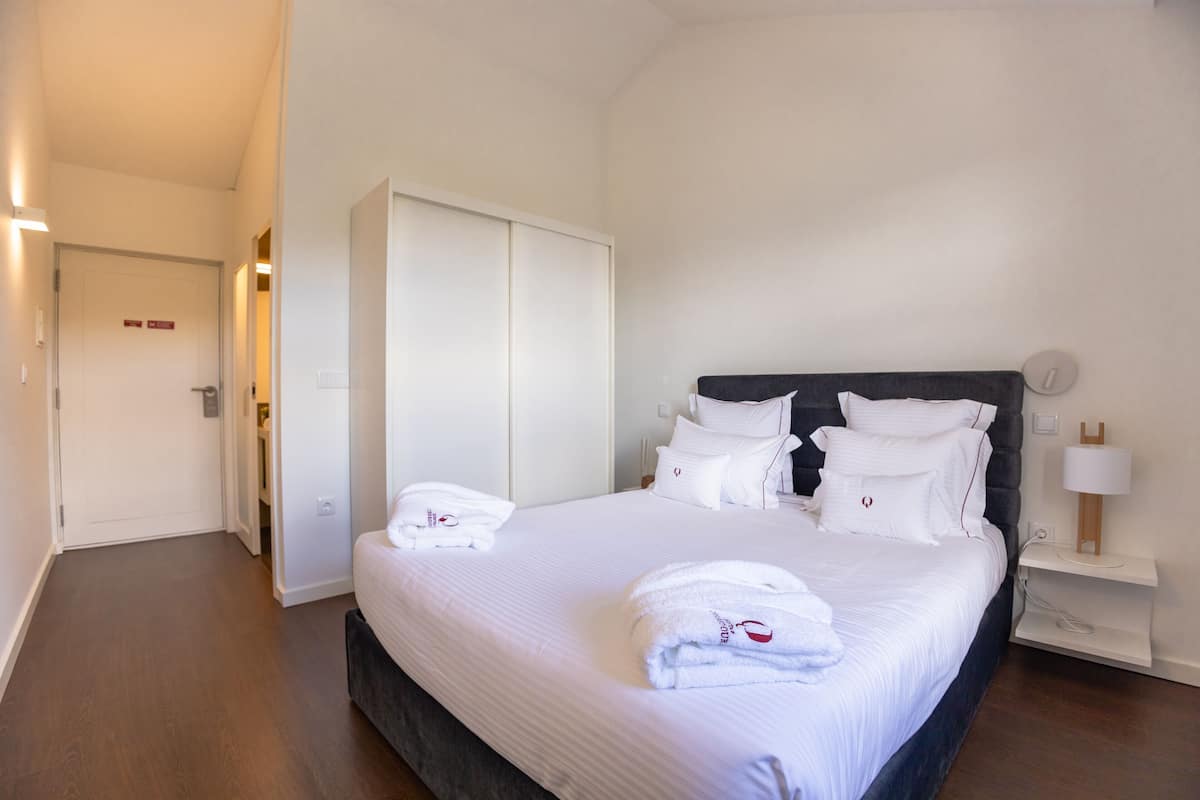 Quarto de hotel com uma cama grande, um guarda-roupa, uma mesa de cabeceira com uma luminária e uma porta que dá para outro quarto.