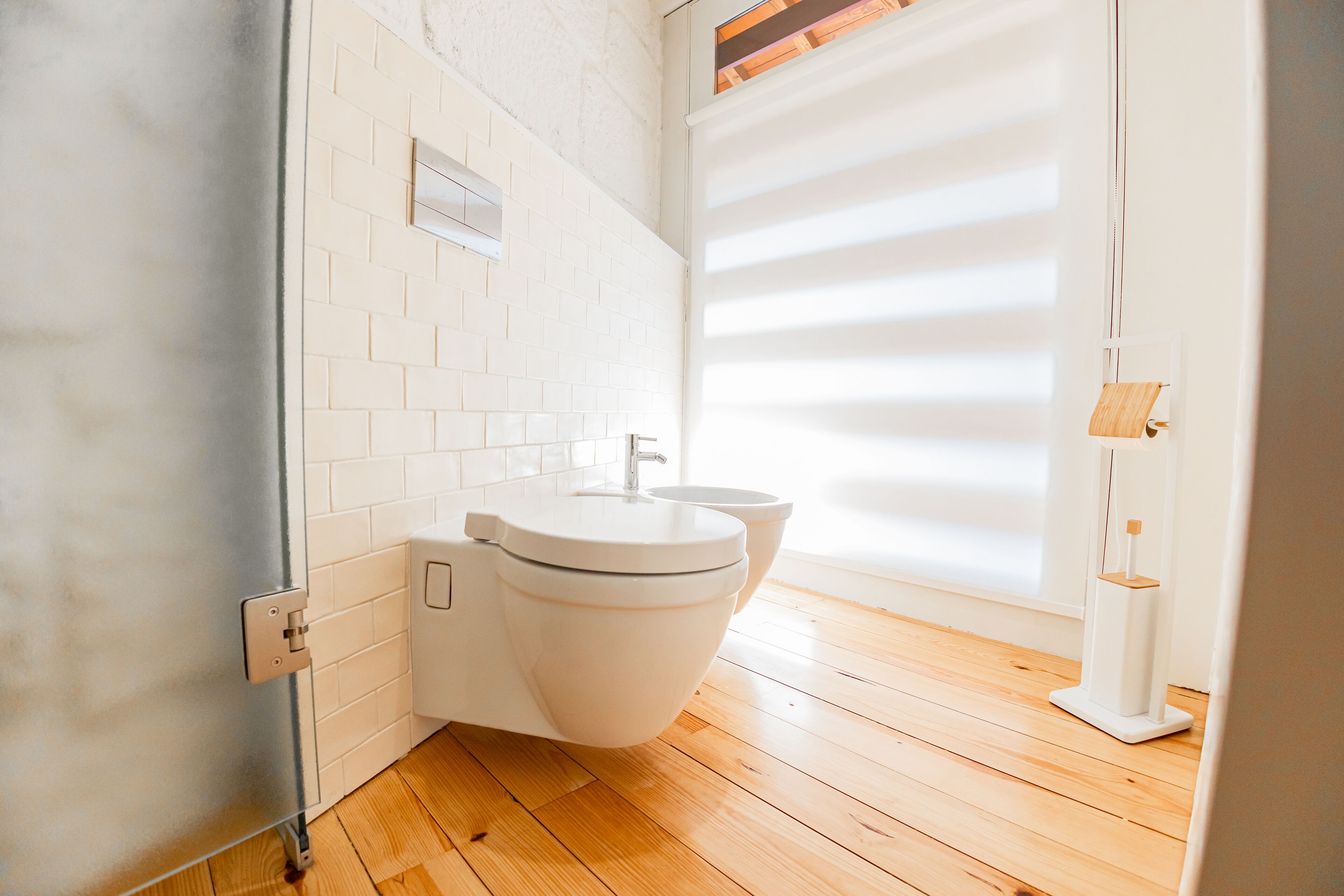 Ein modernes Badezimmer mit weiß gefliesten Wänden, Holzboden und einem Fenster mit Jalousien.
