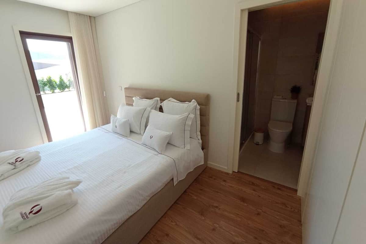 Quarto de hotel com cama, roupa de cama branca, piso de madeira, banheiro visível através de uma porta aberta e uma janela com cortinas.