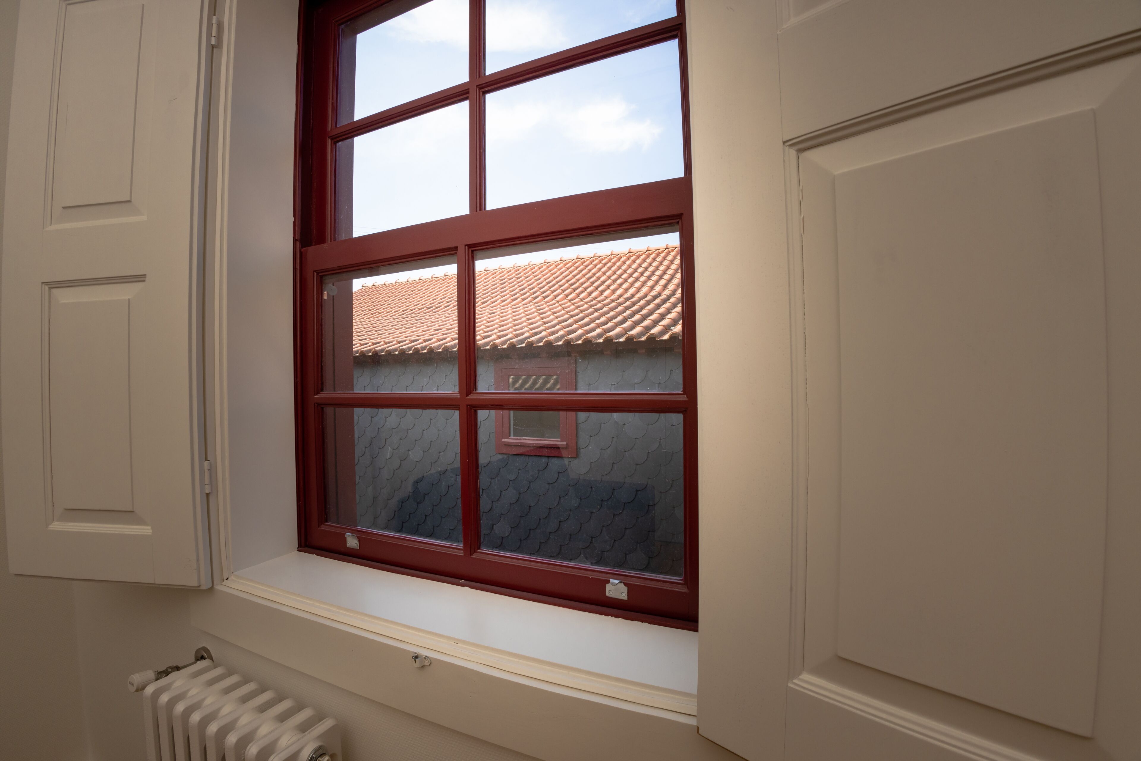 A window with a red frame, a radiator below, and a view of a tiled roof outside.