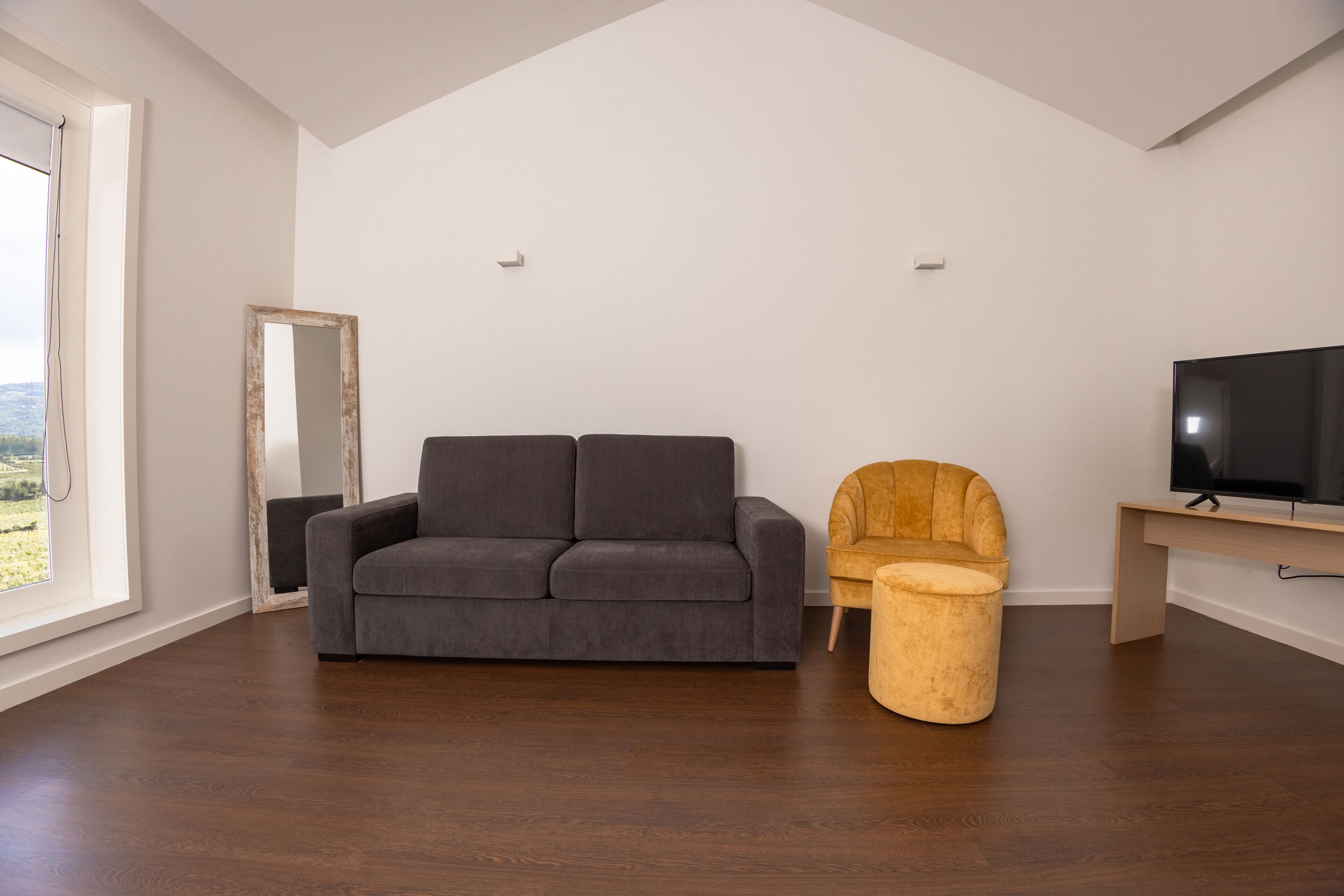 Ein modernes Wohnzimmer mit einem grauen Sofa, einem Holzstuhl, einem runden Hocker und einem Fernseher auf einem Holzboden.
