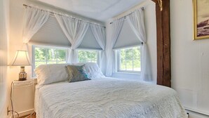 A bedroom with a large bed, two windows with white curtains, a bedside lamp, and a wooden beam on the ceiling.