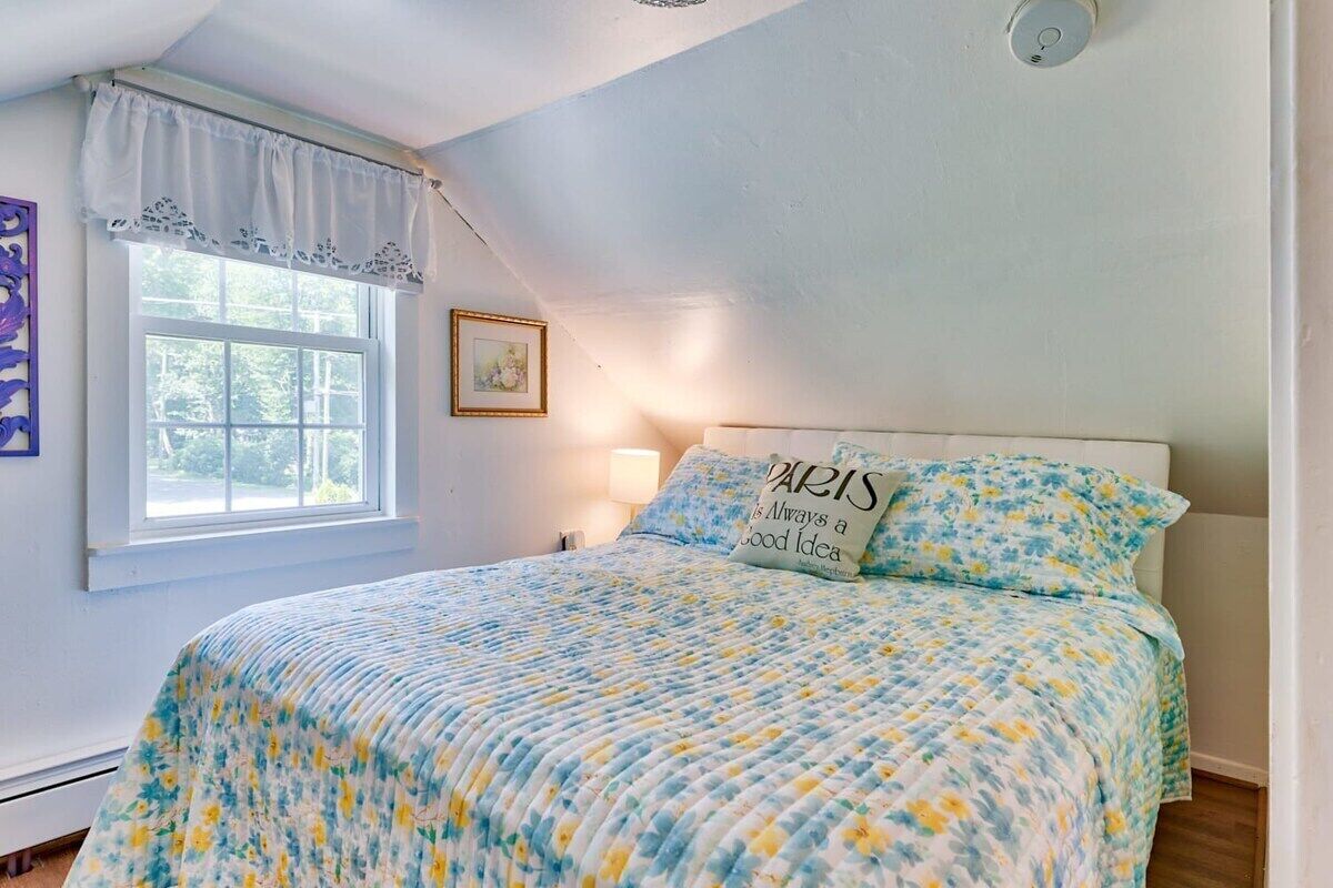 A bedroom with a bed, a window with a curtain, a wall-mounted light, and a framed picture.