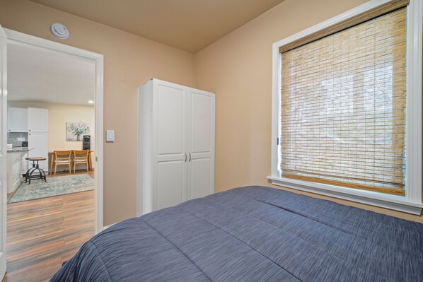 A bedroom with a bed, a wardrobe, a window with blinds, and a view of a kitchen and dining area. - Quaint trendy home just a stroll from the seas (Seaside)