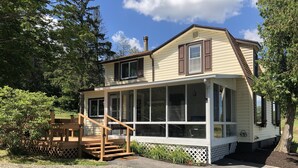 Une maison à deux étages, dotée d’un porche et d’un espace couvert, entourée d’arbres et bordée d’une allée en gravier.
