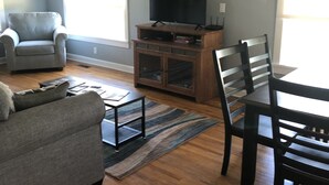 Un salon avec un canapé gris, une table à manger en bois avec des chaises, un téléviseur posé sur un meuble en bois et une fenêtre avec des rideaux.