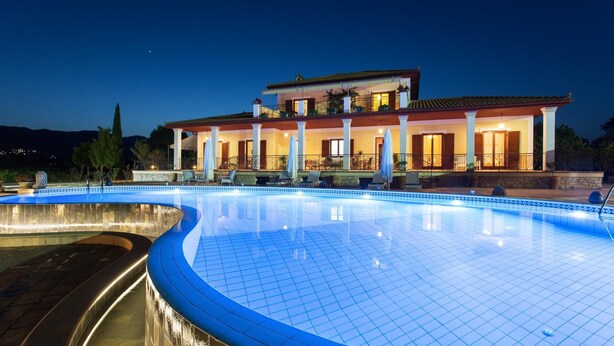 A large, illuminated swimming pool with a curved edge, surrounded by a two-story building with balconies and outdoor seating.