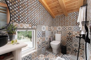 A bathroom with a geometric tile pattern, a toilet, a sink with a window, and a potted plant. - Aneli Villas - Villa Semeli (Zakynthos)